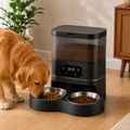 A golden retriever dog is eating from a black automatic pet feeder with two stainless steel bowls in a modern living room setting.