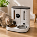 A cat uses a white and black automatic pet feeder and water dispenser on a wooden floor, with a bowl of dry food and water flowing into a second bowl. A potted plant and wooden furniture are visible in the background.